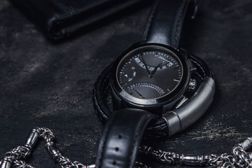 The concept of male style. Black men's watch on a black men's leather bracelet. On the background lies a black wallet. And also a silver chain.