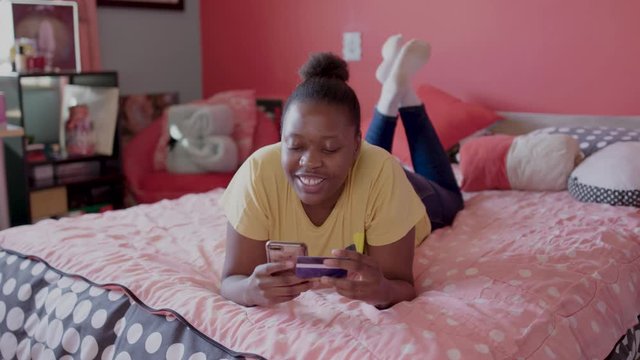 Young black girl makes an online transaction using a smart phone and bank card while lying on her bed. 