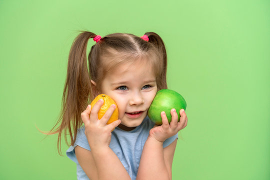 little girl 4 years old in a blue T-shirt on a green background put an apple and a lemon to her face and smiles. pre-school children healthy eating concept