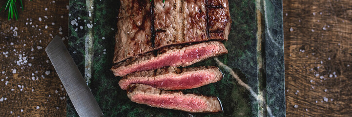 Overhead view of a sliced grilled beef steak on a soapstone with cut, rosemary and coarse salt. Wooden table.