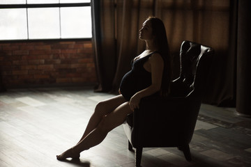 Pregnant women, photoshoot in dark key sit on the armchair
