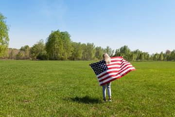 A girl with blond hair and an American flag in her hands behind her back in blue jeans and sneakers runs in a field on green grass against a background of birches.