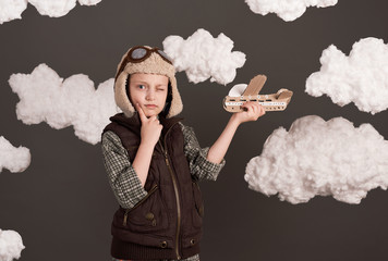 a girl plays with a cardboard airplane and dreams of becoming a pilot, dressed in a retro style jacket and helmet with glasses, clouds of cotton wool, gray background, tinted in brown