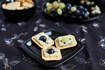 Three blue cheese crackers with grapes are on a black plate.