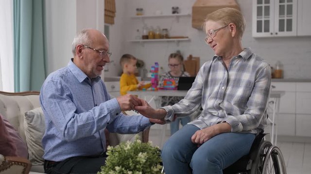 Close-up of loving elderly man taking care of wife with partial paralysis after stroke sitting in wheelchair. Husband helping beloved woman disabled after brain hemorrhage with rehabilitation at home