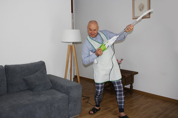 bald man at the age of 55 in home clothes and an apron is dancing happily with a vacuum cleaner, imitating playing the guitar, a cat is sitting on a sofa, a concept of household chores