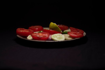 Fresh green cucumber, onions and tomato slices along with green chili and lemons are arranged on a wooden dish in dark copy space background. Food and product photography.