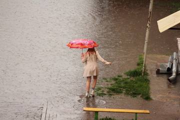 A girl in a short light coat under a colored umbrella stands on the edge of a huge puddle next to a...