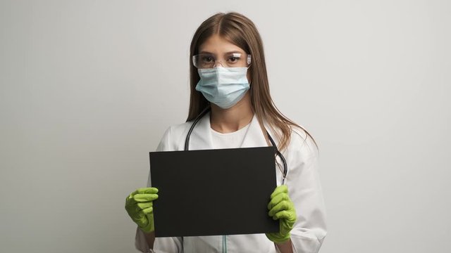 A female doctor in a mask of Caucasian appearance holds a black A4 sheet in her hands
