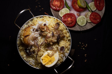 Indian spicy dish Biryani with egg, potato and Chicken in a pot along with fresh salads in dark copy space background. Food and object photography.