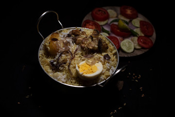 Indian spicy dish Biryani with egg, potato and Chicken in a pot along with fresh salads in dark copy space background. Food and object photography.