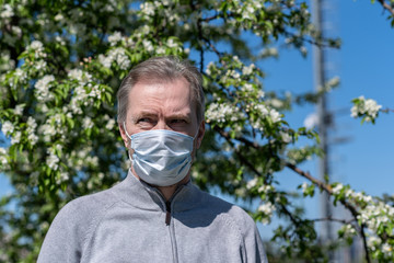 White-haired middle-aged man in a medical mask in a park among flowering trees. Seasonal Allergy. Spring time at sunny day.