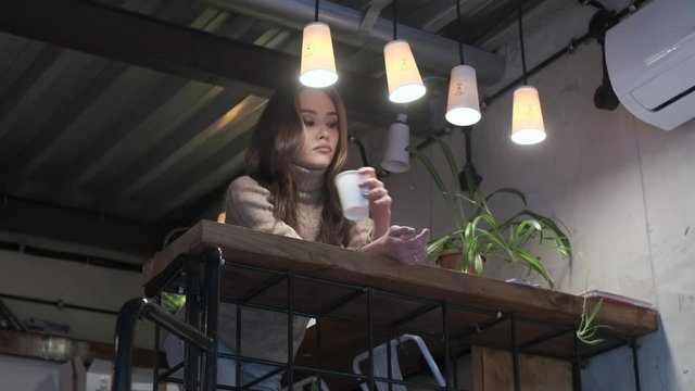 Stylish young asian thoughtful pretty girl wearing beige sweater with long hair sits, holds cup, drinks coffee, warms up and thinks at cozy cafe. Relaxing at restaurant