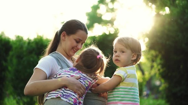 family, children, motherhood, entertainment and people concept - young cute mother of many children with her baby in sling plays with two older kids same age on background of the sunset in the park.