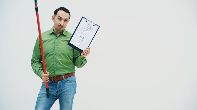 Funny young man standing with the paper holder with word "bisiness" writen on it, looking silly, holding and moving a broomstick.