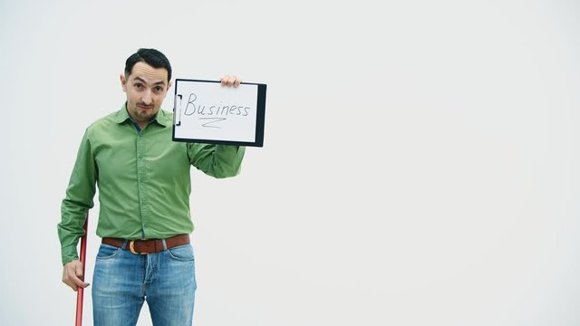 Funny young man standing with the paper holder with word "bisiness" writen on it, looking silly, holding and moving a broomstick, then throw it like a spear.