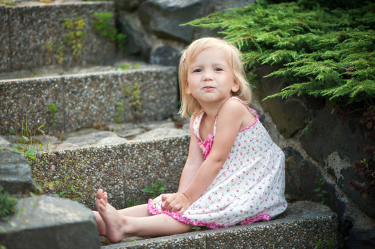 A little girl in a dress sits on the stone steps in the garden. Bare feet, smile.