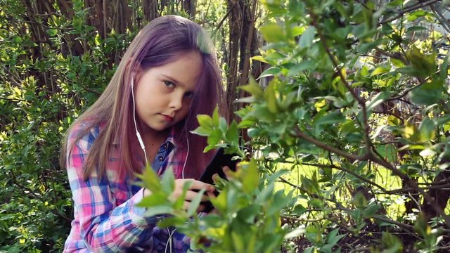 little beautiful girl plays on the phone and listens to music on the background of foliage shrubs and trees.