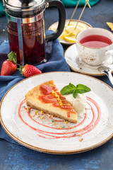 Cheesecake slice with strawberry sauce and ice cream on white plate with cup of tea