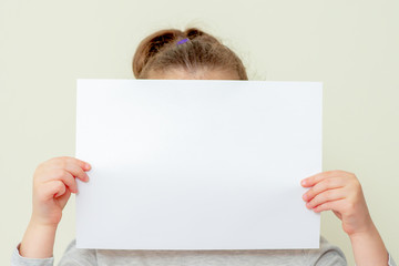 Child's hands holding empty sheet of a paper covering her face on white background. Mockup sheet of paper in child's hands.