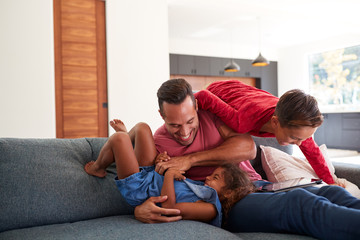 Father Using Digital Tablet At Home Tickling Son And Daughter On Sofa