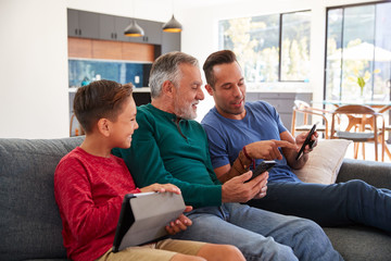 Multi-Generation Male Hispanic Family Sitting On Sofa At Home Using Mobile Phones And Digital Tablet