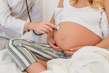 Doctor in clinic listening with phonendoscope belly of pregnant blonde woman . Future mother with naked belly have prenatal consultation. Concept of professional healthcare during pregnancy