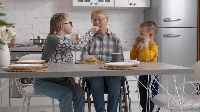 Loving grandchildren helping paralyzed grandmother having breakfast in home kitchen. Granddaughter feeding beloved handicapped granny with oatmeal from spoon while grandson wiping her lips with napkin