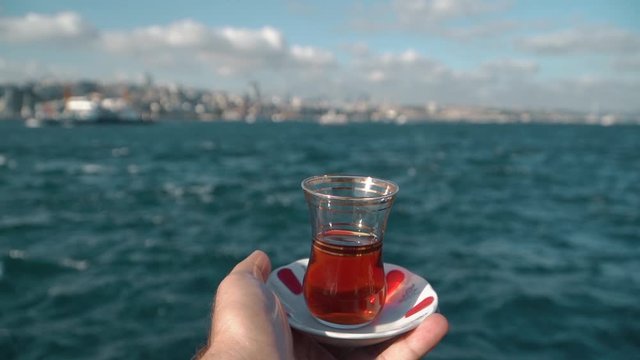 Traditional black Turkish tea in a glass on a plate in the man hands with a view of the Bosphorus and Istanbul background. Travel to Turkey. Soft focus and beautiful bokeh. Slow motion shutting.