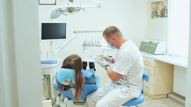 The girl in the chair at the dentists appointment. The doctor in the office shows the patient a layout of the dental jaw.