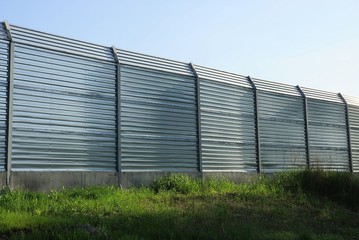 high gray metal fence wall in green grass on the street against a blue sky