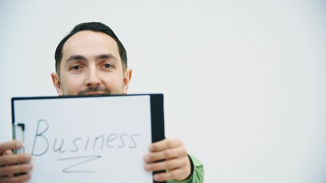 Funny young man standing holding the paper holder with word "bisiness" writen on it, showing it at the camera, raising eyebrows.