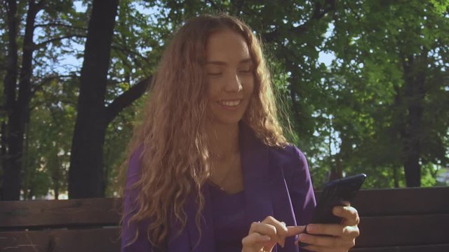 Smiling girl places order in online store and makes purchase payment. Remote Shoping. Curly long haired woman in suit without makeup looks at smartphone and watching news feed. Communication. Vlogger