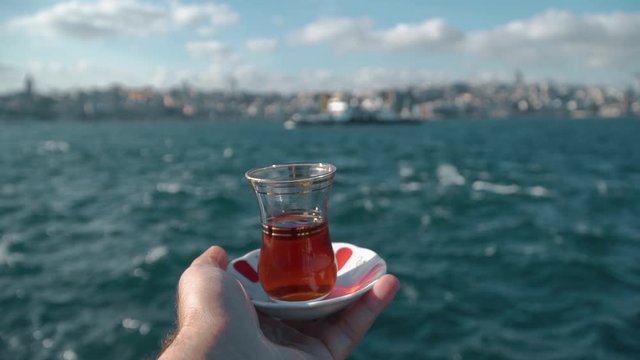 Traditional black Turkish tea in a glass on a plate in the man hands with a view of the Bosphorus and Istanbul background. Travel to Turkey. Soft focus and beautiful bokeh. Slow motion shutting. Part2