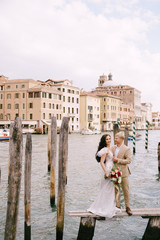 Fototapeta na wymiar Italy wedding in Venice. The bride and groom are standing on a wooden pier for boats and gondolas, near the Striped green and white mooring poles, against backdrop of facades of Grand Canal buildings.
