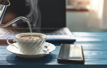 A white cup of coffee with hot smoke in stainless pot, Morning coffee time with notebook computer and work equipment, Normal practice by working at home during covid-19 outbreak around the world.