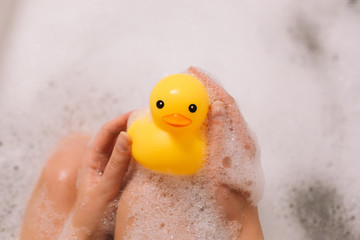 Girl is holding yellow rubber duck in the bath with bubble foam