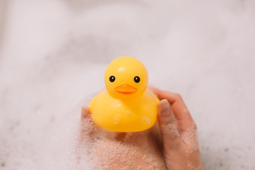 Girl is holding yellow rubber duck in the bath with bubble foam