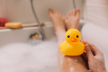 Girl is holding on her legs yellow rubber duck in the bath with bubble foam