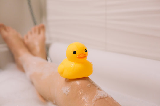 Girl is holding on her legs yellow rubber duck in the bath with bubble foam