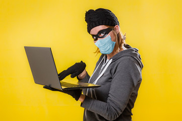 A hacker in a medical mask is holding a laptop. Yellow background. The concept of cybercrime growth in the context of the coronavirus crisis