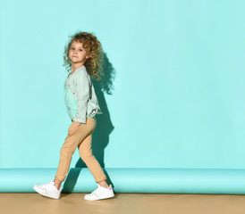 Little kid in denim jacket, beige pants and white sneakers. She smiling, hands in pockets, posing sideways on turquoise background