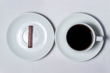 One chocolate tube on a white saucer and a cup of coffee on a white tablecloth. The saucer and cup are also white