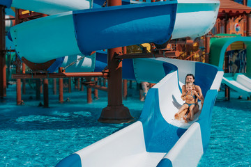 Little girl in swimsuit goes down the blue slides to the pool, Mom and daughter play and swim in...