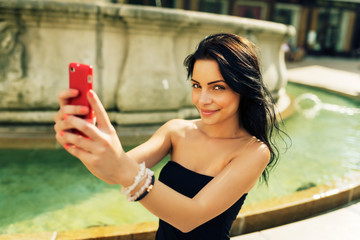 Beautiful girl smiling and taking selfie. Young pretty woman having fun and taking photo with red smartphone in front of water fountain in historical part of Bratislava city in hot summer afternoon.