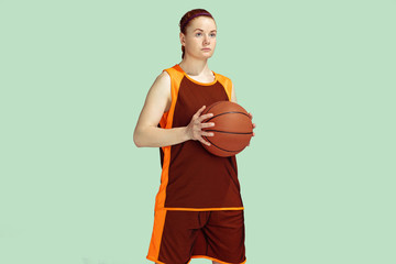 Young caucasian female basketball player confident holding ball, posing isolated on mint colored background. Concept of sport, movement, energy and dynamic, healthy lifestyle. Training, practicing.