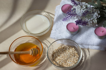 Home Spa skin care ingredients. Glass jars of oatmeal and yellow honey, a white towel for the bathroom. There is a lavender flower in the background. Side view. Sunlight. Dripping honey.