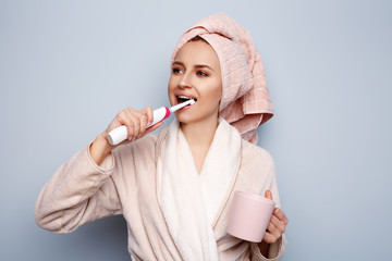 Woman in pink bathrobe with towel on head brushing teeth with electric toothbrush, holding cup of coffee or tea, morning routine after shower. White teeth and health care. Cheerful ready for new day