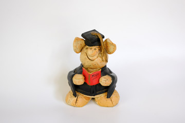 Figure of a ceramic pig wearing a graduation holding a red book with the cover text "MUM" 