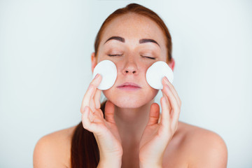 portrait of redheaded young woman looking natural wiping her face with cotton pads standing on...